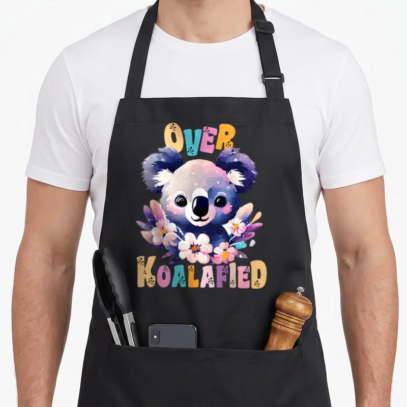 Koalafied floral apron - Cartoon koala head with pink purple blue orange flowers and arched OVER KOALAFIED text — shown on a black cotton canvas apron against a white background