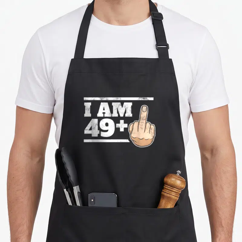 Milestone joke apron - I AM 49+ text with a middle finger hand in distressed white and skin tone — shown on a black cotton canvas apron against a white background