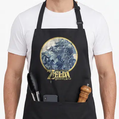 Breath rock apron - Four blue watercolor characters in gold circle with Zelda text and sword — shown on a black cotton canvas apron against a white background