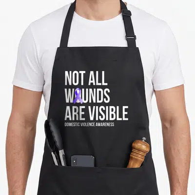 Visible domestic aprons - White text Not All Wounds Are Visible with purple ribbon inside W on black background — shown on a black cotton canvas apron against a white background