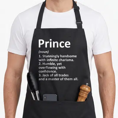Prince definition birthday apron - White dictionary definition text reading Prince noun with three descriptive points — shown on a black cotton canvas apron against a white background