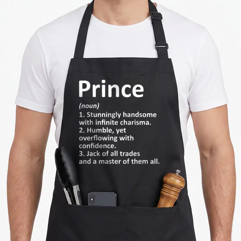Prince definition birthday apron - White dictionary definition text reading Prince noun with three descriptive points — shown on a black cotton canvas apron against a white background