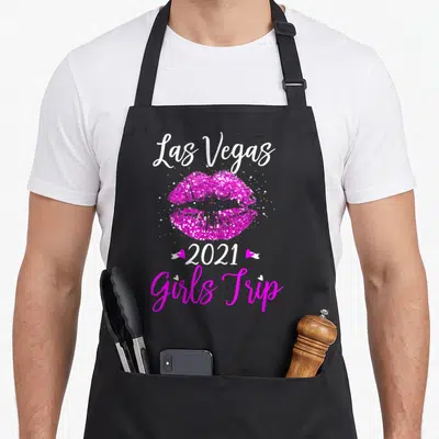 Trip vegas girls apron - White Las Vegas text, pink glitter lips, 2021 with two hearts, pink Girls Trip script — shown on a black cotton canvas apron against a white background