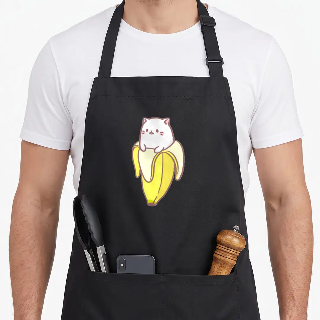 Cat banana apron - White cartoon cat sitting inside a peeled yellow banana with pink ears — shown on a black cotton canvas apron against a white background