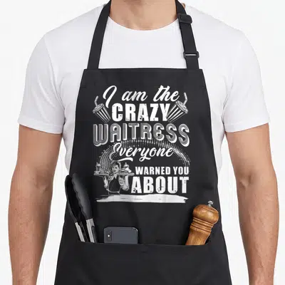 Waitress novelty apron - White stacked text reading I am the crazy waitress everyone warned you about with server — shown on a black cotton canvas apron against a white background