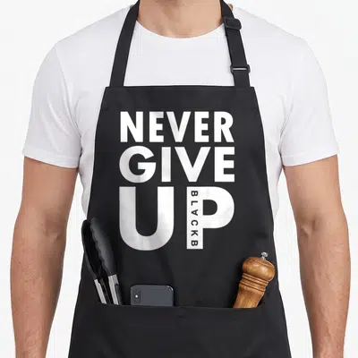 Give golf fan apron - White NEVER GIVE UP text stack with BLACKB written vertically inside the letter P — shown on a black cotton canvas apron against a white background