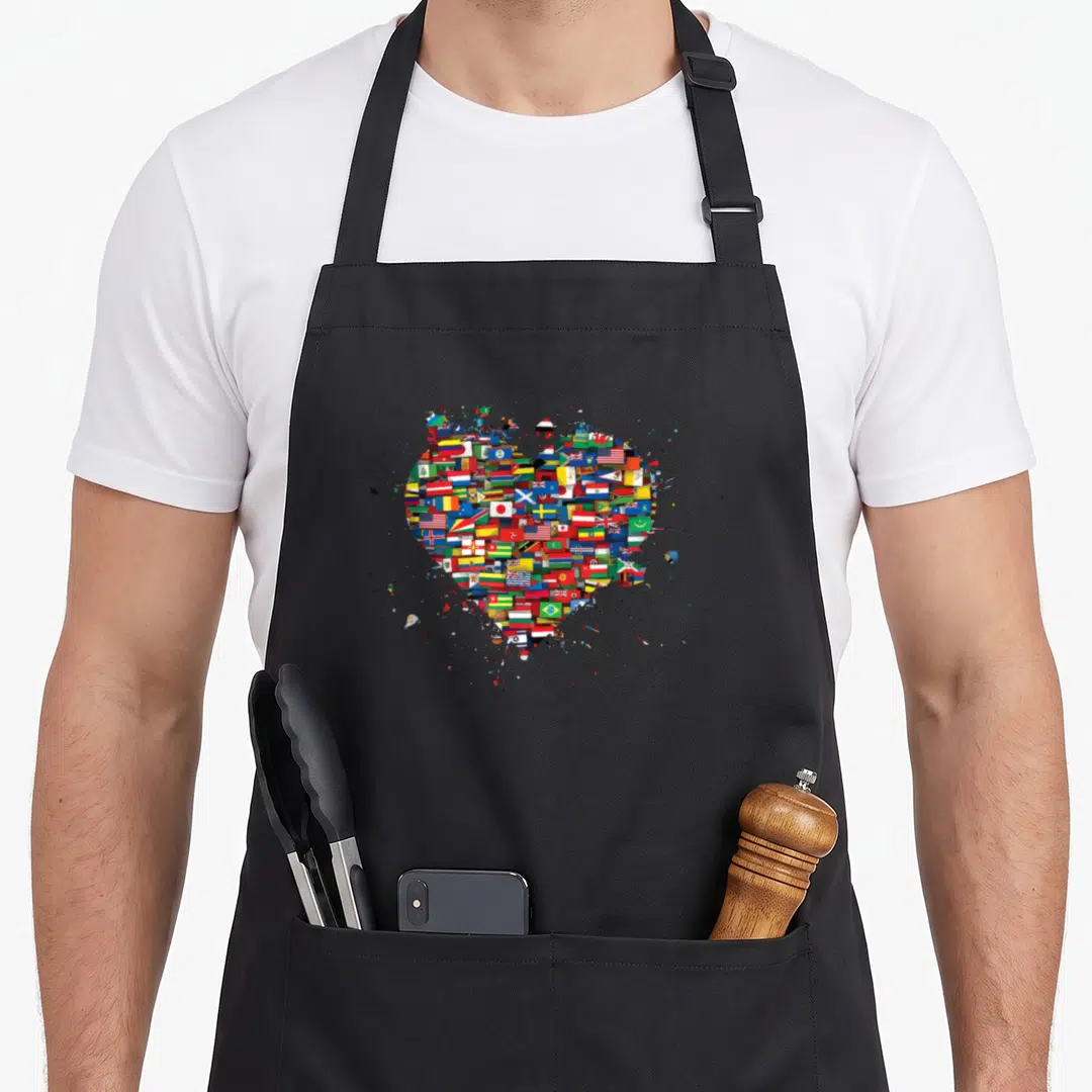 Funny flag country apron - Heart shape formed by small multicolored international flags surrounded by paint splatters — shown on a black cotton canvas apron against a white background