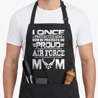 Air show mama apron - Distressed white text I once protected him now he protects me proud air force mom with wings — shown on a black cotton canvas apron against a white background