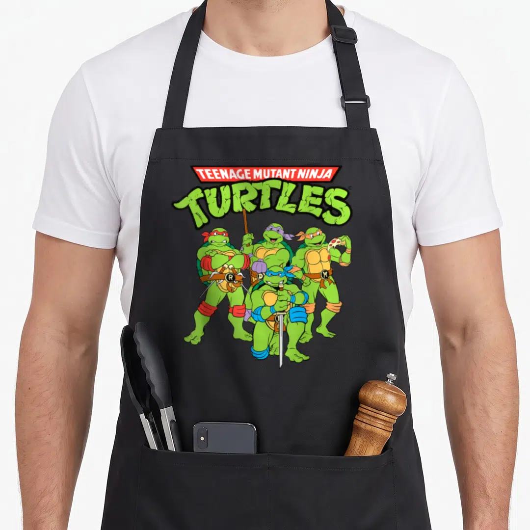 Cute teenage cooking aprons - Stacked TMNT logo above four green turtles wearing red blue purple and yellow masks — shown on a black cotton canvas apron against a white background