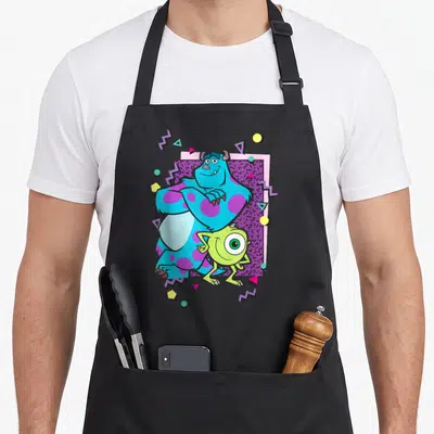 Mike 90s apron - Sulley and Mike centered on purple patterned rectangle with colorful geometric shapes and zigzags — shown on a black cotton canvas apron against a white background
