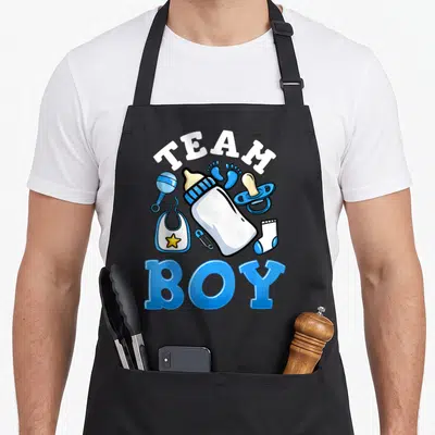 Vintage gender reveal apron - TEAM BOY text with blue baby bottle, bib, and footprints in cartoon style — shown on a black cotton canvas apron against a white background