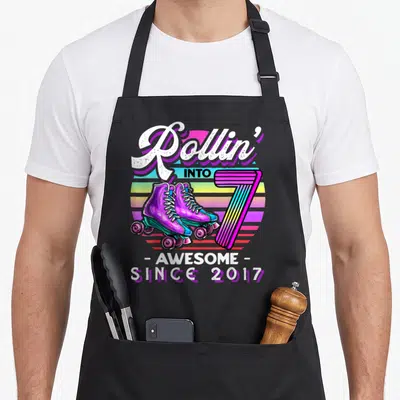 7th rollin birthday apron - Retro roller skates next to striped number seven and rainbow sunburst background — shown on a black cotton canvas apron against a white background