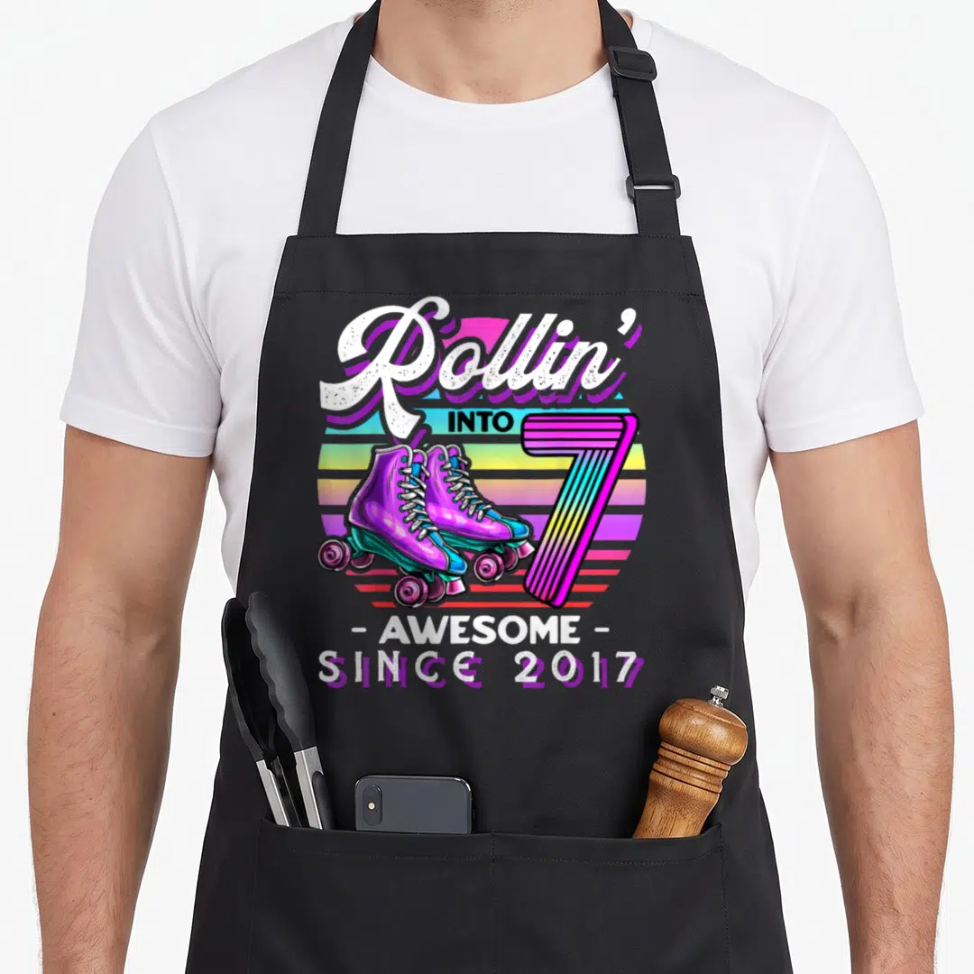 7th rollin birthday apron - Retro roller skates next to striped number seven and rainbow sunburst background — shown on a black cotton canvas apron against a white background