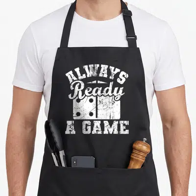 Ready game aprons - Distressed white Always Ready A Game text over a domino tile on black fabric — shown on a black cotton canvas apron against a white background