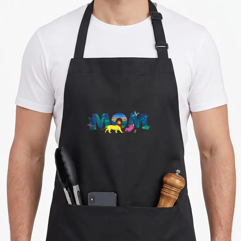 Pal mother's day aprons - MOM letters filled with cartoon lion warthog bird and jungle scene in blue yellow green pink — shown on a black cotton canvas apron against a white background