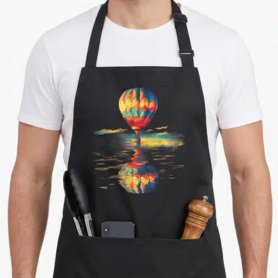 Ballooning balloon apron - Watercolor hot air balloon with blue yellow red orange segments reflecting on water surface — shown on a black cotton canvas apron against a white background