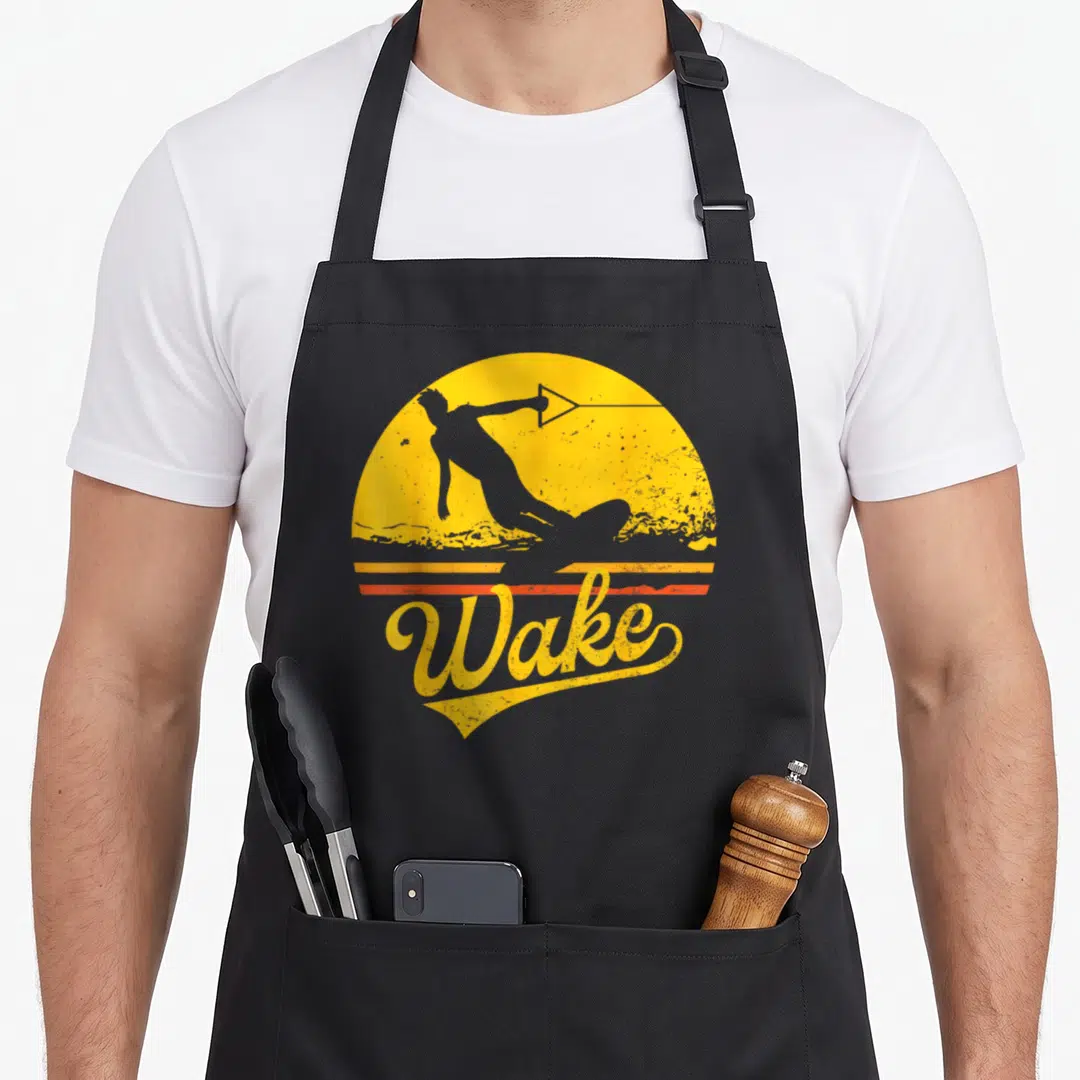 Sunset boarding apron - Retro wakeboarder silhouette on yellow sun with grey water and orange stripes below the word — shown on a black cotton canvas apron against a white background