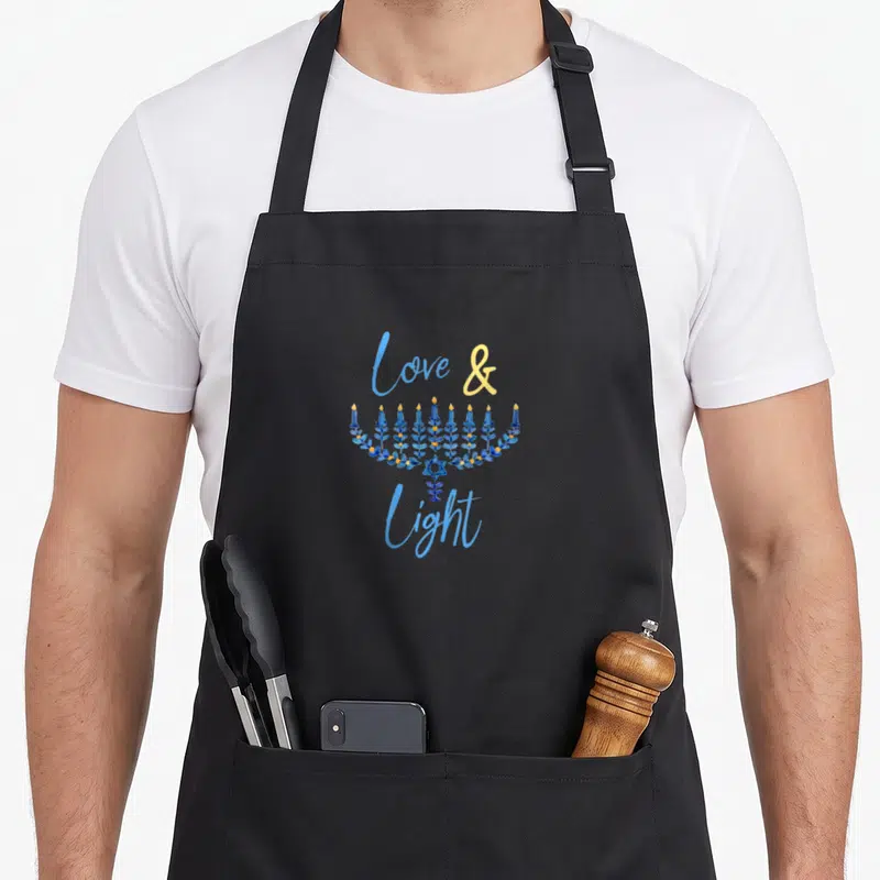 Blue hanukkah apron - Cartoon menorah in blue and yellow with star of David and love and light text — shown on a black cotton canvas apron against a white background