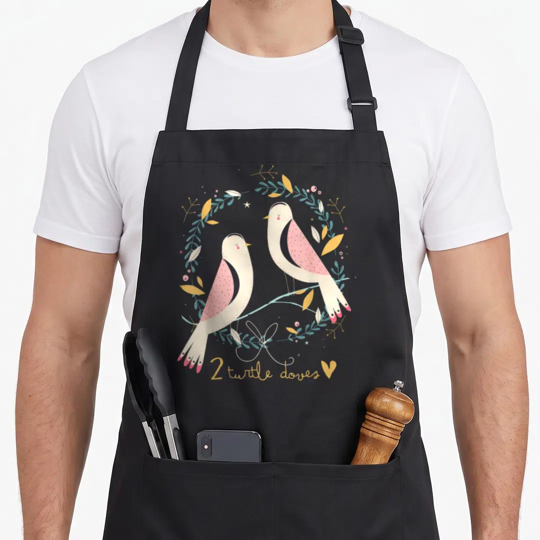 Dove christmas apron - Two cartoon birds on a branch inside a wreath with 2 turtle doves text and a heart — shown on a black cotton canvas apron against a white background