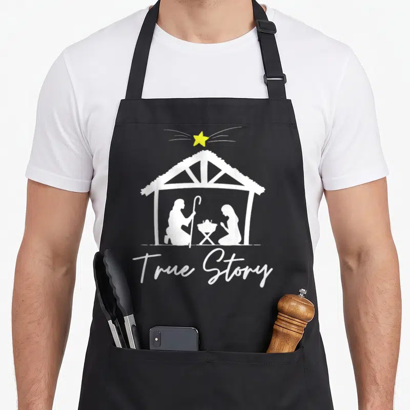 Story jesus womens christmas apron - White stable silhouette with three figures and yellow star above True Story text — shown on a black cotton canvas apron against a white background