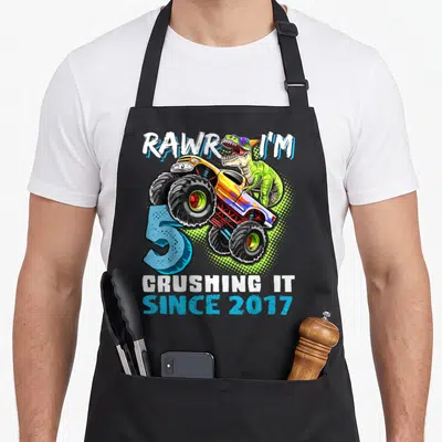 Rawr monster truck apron - Dinosaur wearing sunglasses driving monster truck with blue number 5 and celebratory text — shown on a black cotton canvas apron against a white background