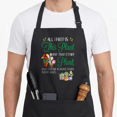 Funny plant gardener apron - Cartoon potted plants in green red yellow and white with stacked text ALL I NEED IS This — shown on a black cotton canvas apron against a white background