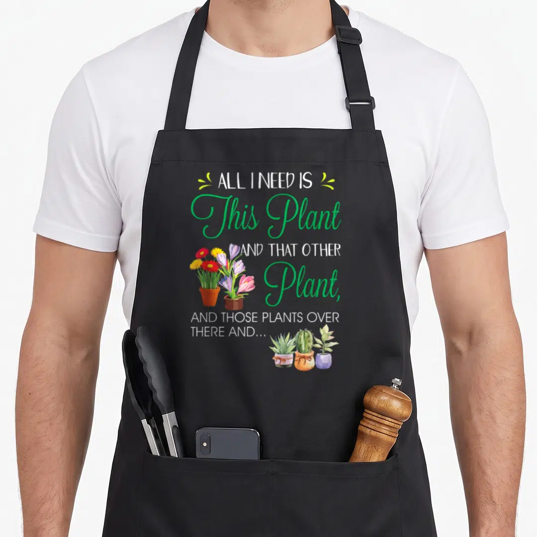 Funny plant gardener apron - Cartoon potted plants in green red yellow and white with stacked text ALL I NEED IS This — shown on a black cotton canvas apron against a white background