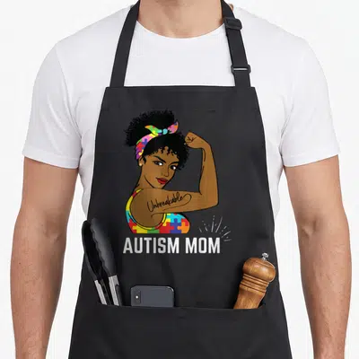 Awareness mother's apron - Cartoon woman flexing arm with Unbreakable tattoo and Autism Mom text below — shown on a black cotton canvas apron against a white background