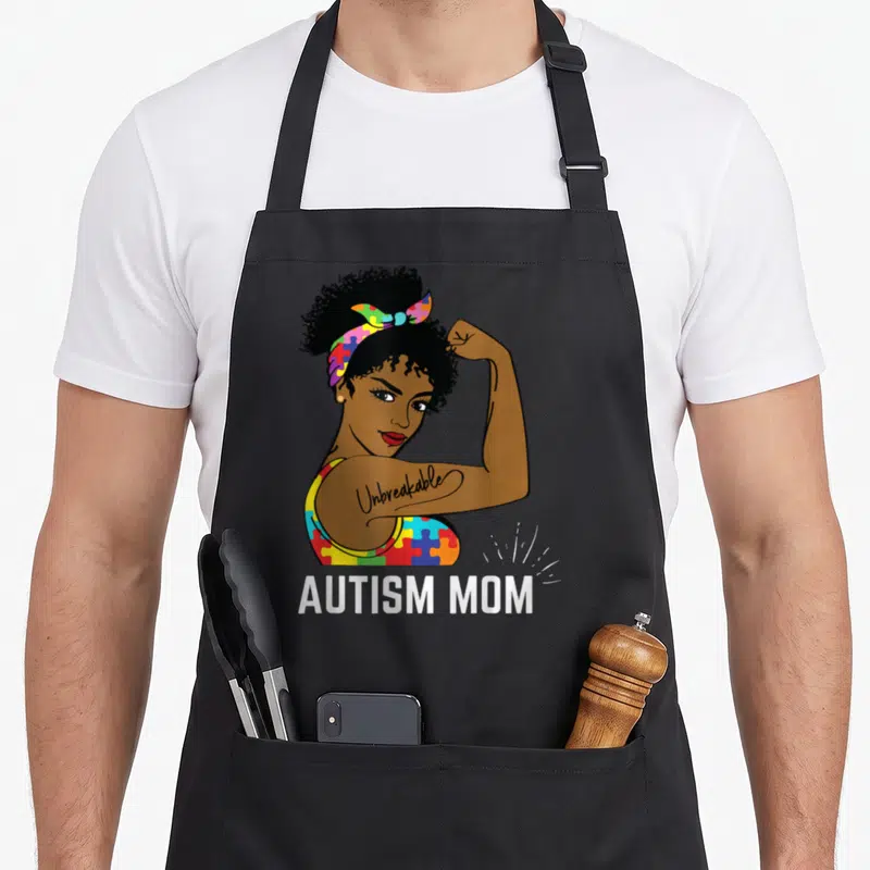 Awareness mother's apron - Cartoon woman flexing arm with Unbreakable tattoo and Autism Mom text below — shown on a black cotton canvas apron against a white background