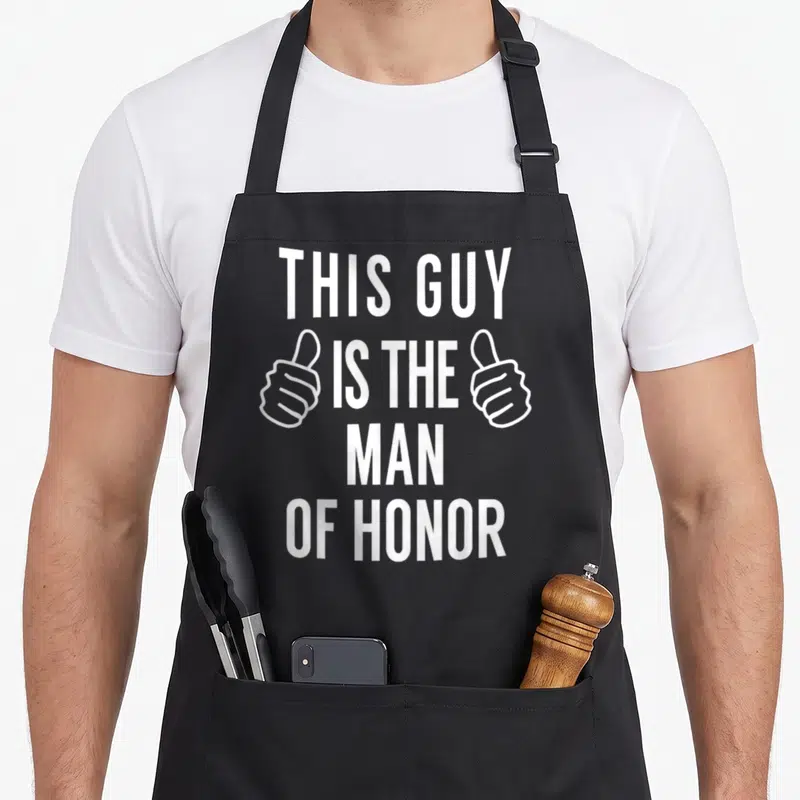Funny your honor apron - White stacked text THIS GUY IS THE MAN OF HONOR with two thumbs up icons — shown on a black cotton canvas apron against a white background