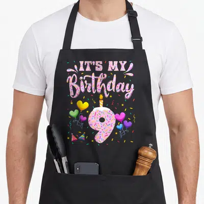 Doughnut birthday apron - IT'S MY Birthday text above frosted number 9 donut with heart balloons and confetti — shown on a black cotton canvas apron against a white background