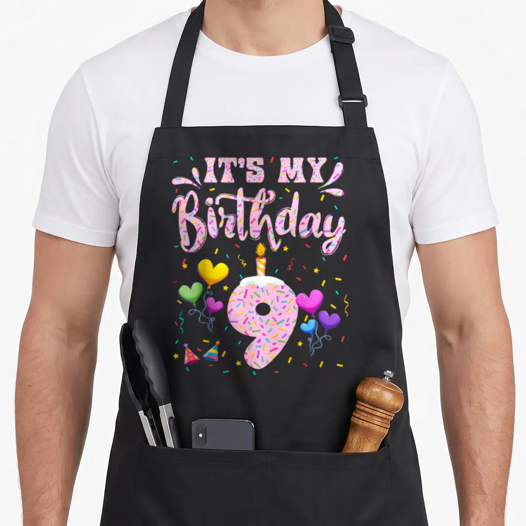 Doughnut birthday apron - IT'S MY Birthday text above frosted number 9 donut with heart balloons and confetti — shown on a black cotton canvas apron against a white background