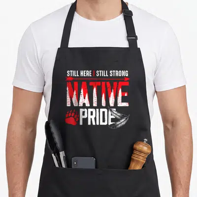 Indigenous native american aprons - Red and white text Still Here Still Strong Native Pride with arrow bear paw — shown on a black cotton canvas apron against a white background