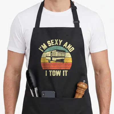 Camper tow trailer apron - Retro striped sun with camper trailer and text I'M SEXY AND I TOW IT — shown on a black cotton canvas apron against a white background