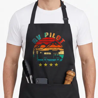 Pilot camping apron - Retro RV PILOT text over sunset with RV, mountains, and four stars in red orange yellow blue grey — shown on a black cotton canvas apron against a white background