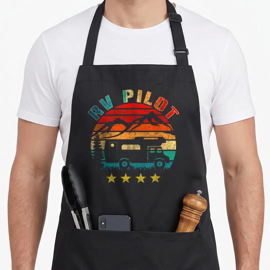 Pilot camping apron - Retro RV PILOT text over sunset with RV, mountains, and four stars in red orange yellow blue grey — shown on a black cotton canvas apron against a white background