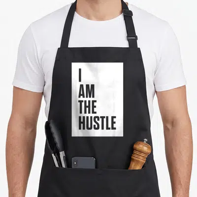 Cute hustle apron - Grey stacked text reading I AM THE HUSTLE on white fabric — shown on a black cotton canvas apron against a white background