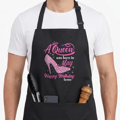 Cute may birthday apron - Pink rhinestone high heel shoe with crown and A Queen was born in May text surrounded — shown on a black cotton canvas apron against a white background