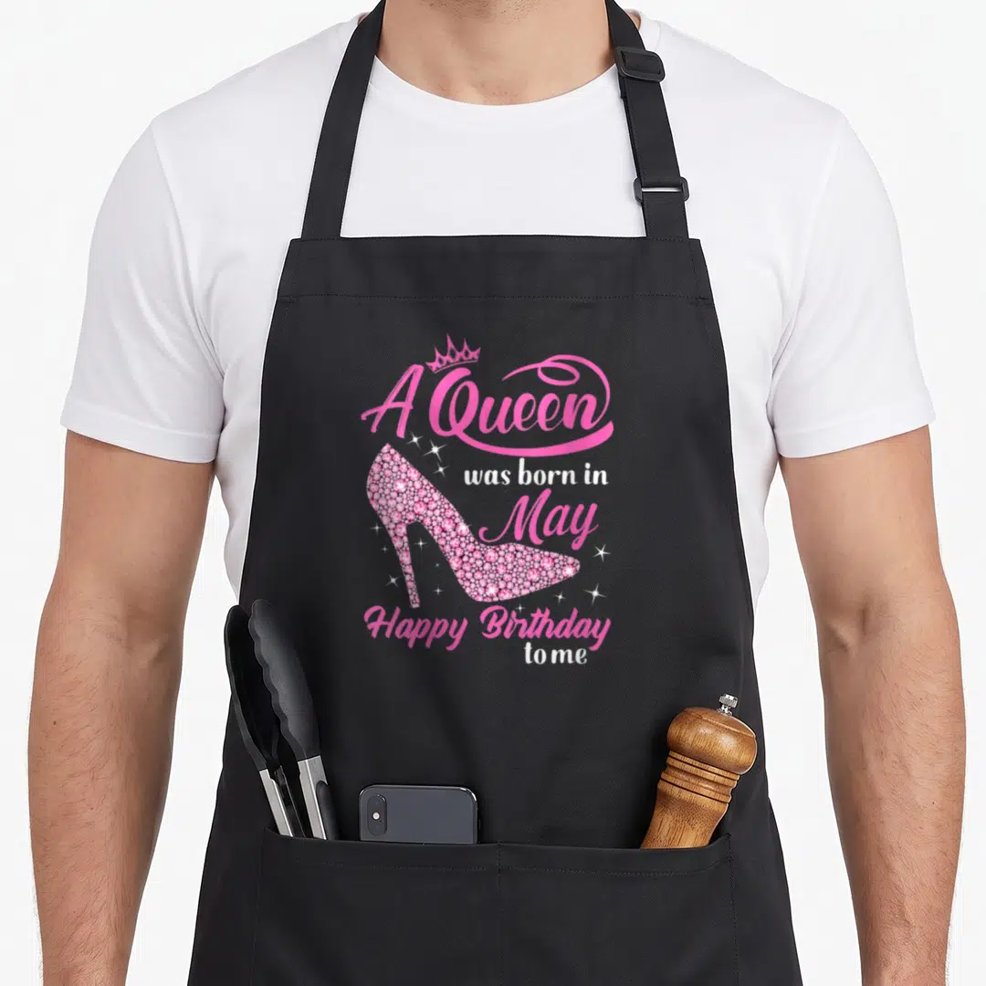 Cute may birthday apron - Pink rhinestone high heel shoe with crown and A Queen was born in May text surrounded — shown on a black cotton canvas apron against a white background