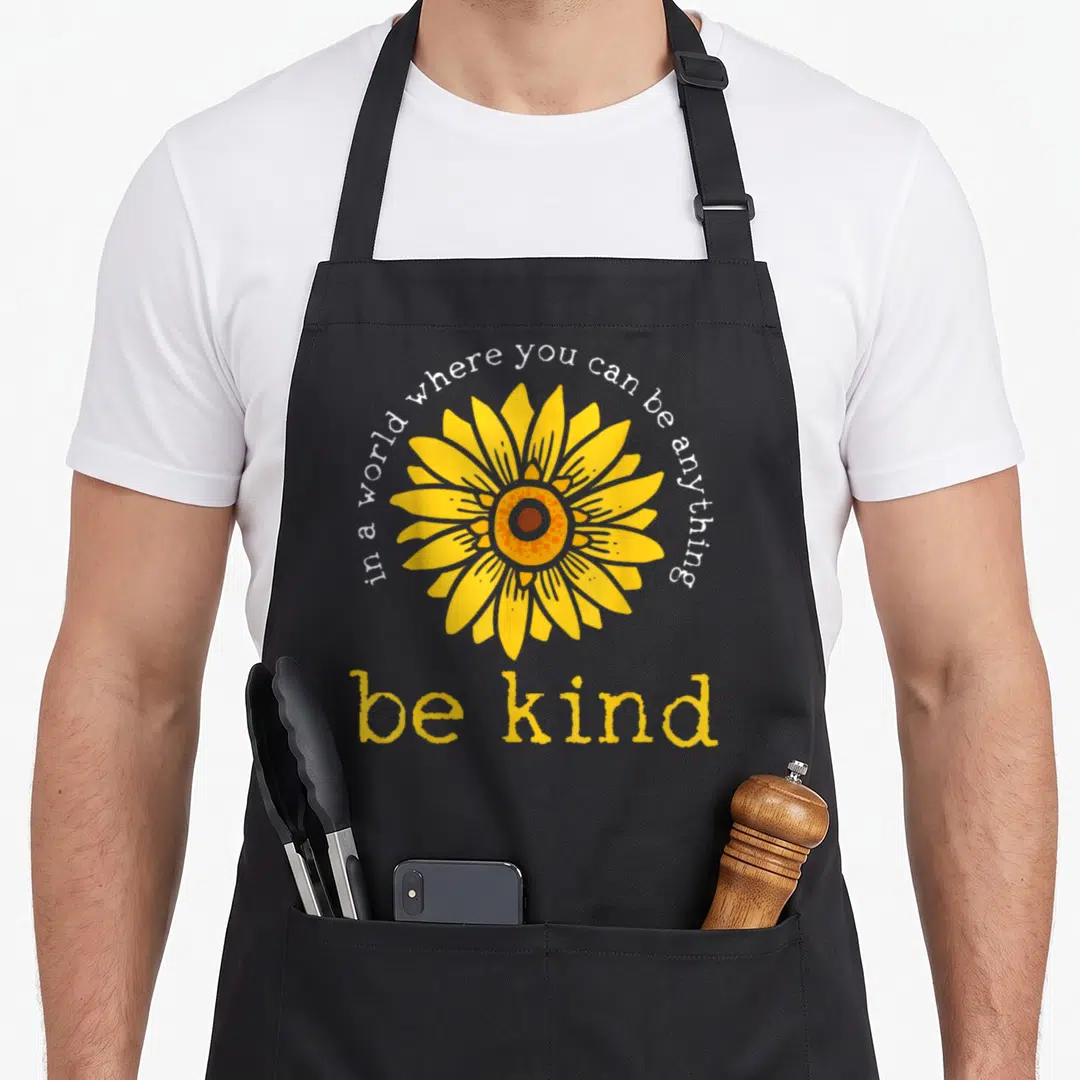 Anything im jay apron - Cartoon sunflower with yellow petals and text in a world where you can be anything be kind — shown on a black cotton canvas apron against a white background