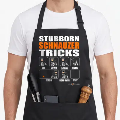 Stubborn schnauzer apron - Cartoon schnauzer grid with Sit Down Shake Come Fetch Roll Over Stay labels — shown on a black cotton canvas apron against a white background