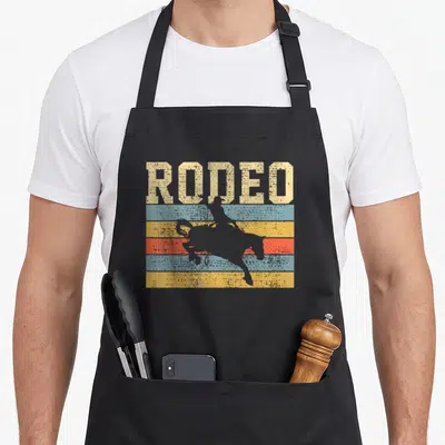 Riding country apron - Distressed RODEO text over grey bull rider silhouette on blue yellow and red stripes — shown on a black cotton canvas apron against a white background