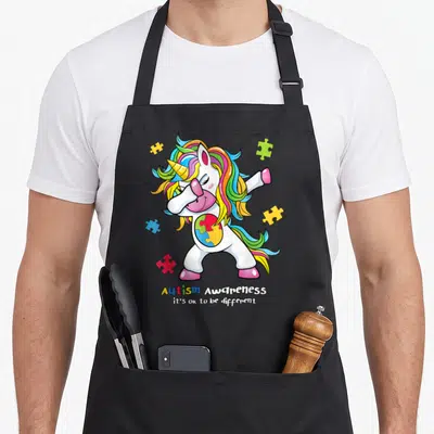 Funny different autism awareness apron - Dabbing unicorn with rainbow mane and puzzle heart chest surrounded by four — shown on a black cotton canvas apron against a white background