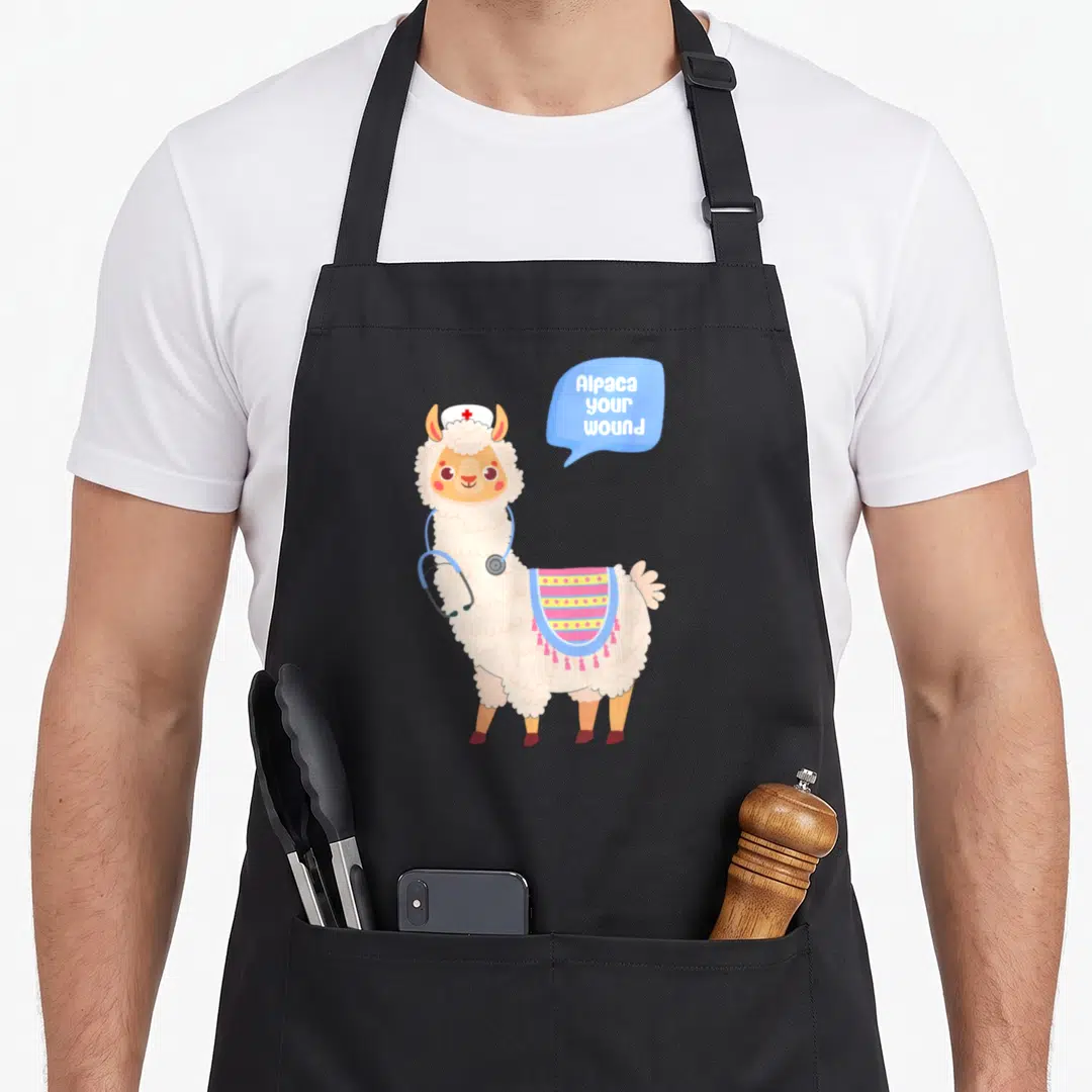 Wound alpaca apron - Cartoon alpaca in nurse hat with stethoscope and speech bubble text — shown on a black cotton canvas apron against a white background