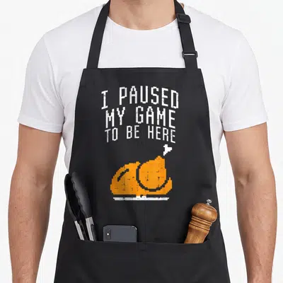 Paused turkey apron - Pixelated orange turkey and white cursor below stacked white text in 8-bit font — shown on a black cotton canvas apron against a white background