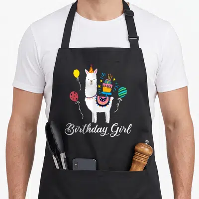 Animal alpaca apron - Cartoon white llama wearing party hat and saddle with cake, balloons, and Birthday Girl text — shown on a black cotton canvas apron against a white background