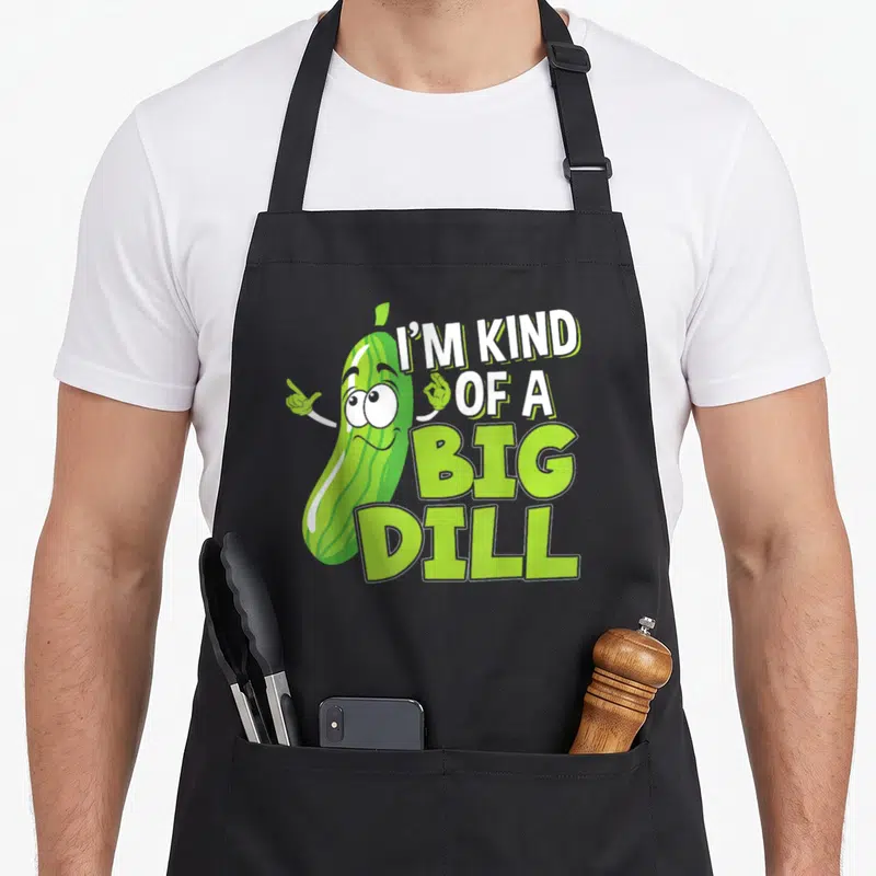 Vegan pickle apron - Cartoon green pickle character pointing next to stacked text reading I'M KIND OF A BIG DILL — shown on a black cotton canvas apron against a white background