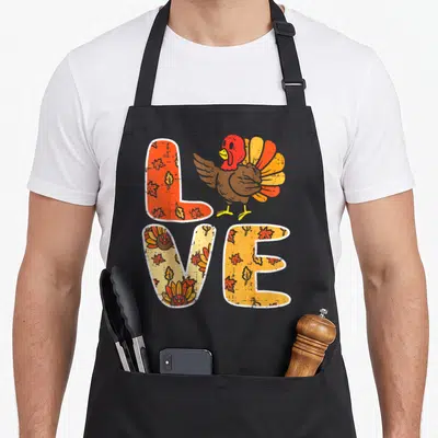 Turkey leave thanksgiving apron - LOVE text filled with leaves and small turkeys with a large cartoon turkey standing — shown on a black cotton canvas apron against a white background
