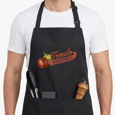 Whisperer tomato apron - Retro red and yellow tomato illustration with whisperer text on black bib — shown on a black cotton canvas apron against a white background