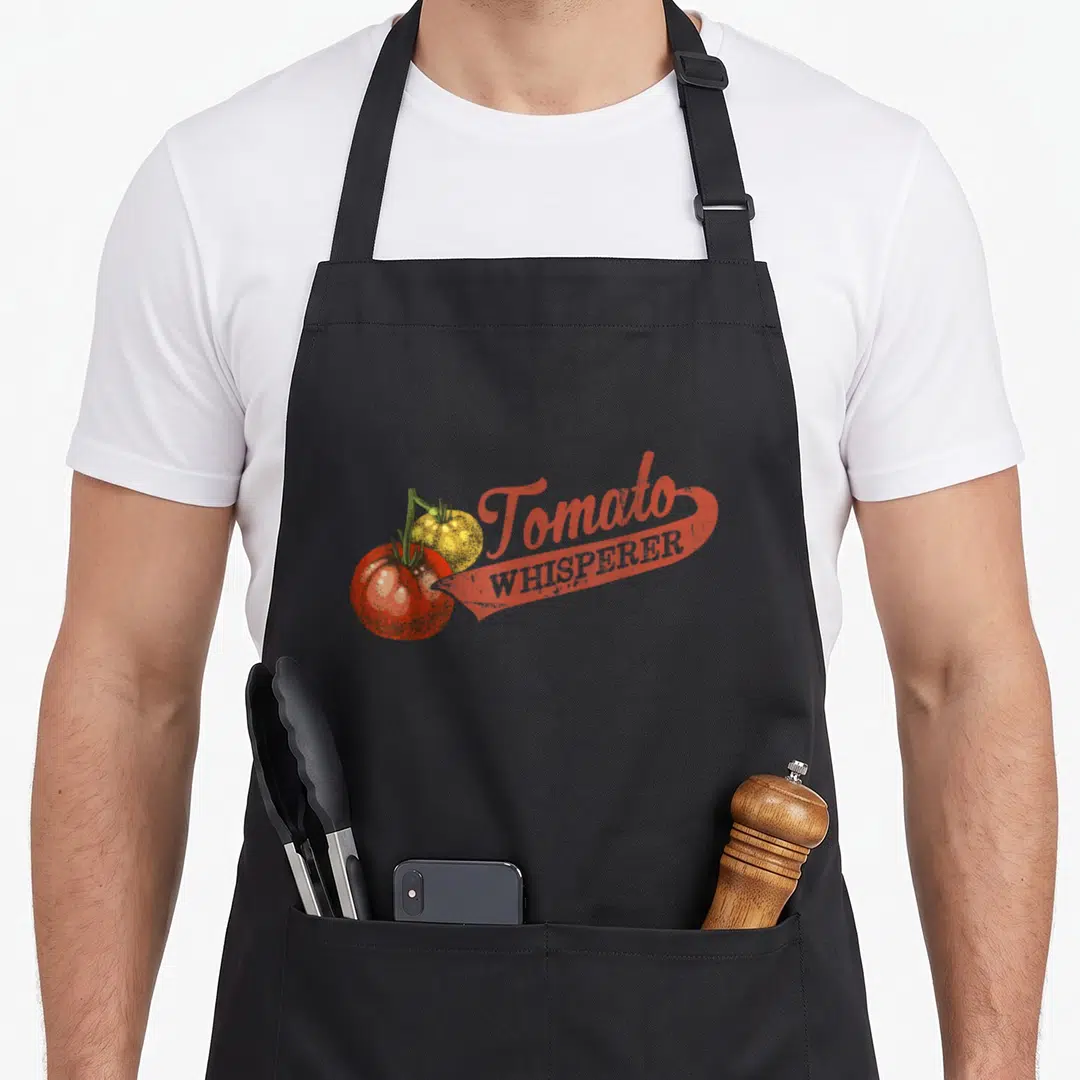Whisperer tomato apron - Retro red and yellow tomato illustration with whisperer text on black bib — shown on a black cotton canvas apron against a white background